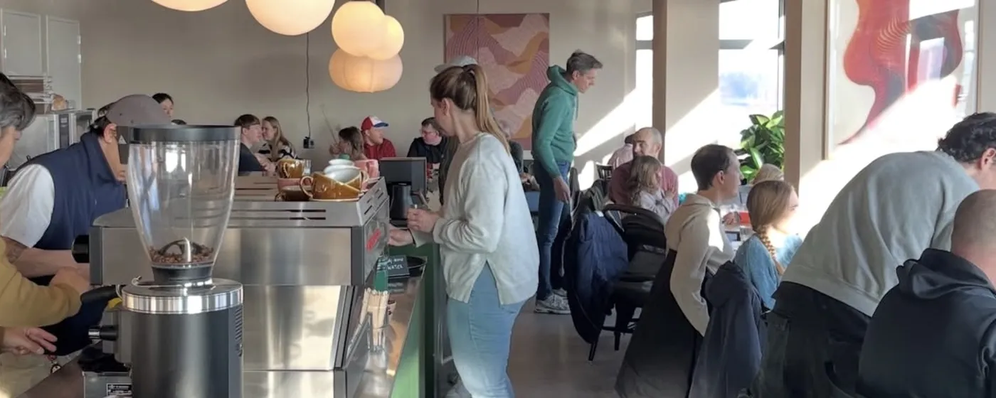 Volle zaal bij KOMU: gasten aan de ramen, iemand bestelt aan de groene bar en de bollampen hangen boven het espressostation